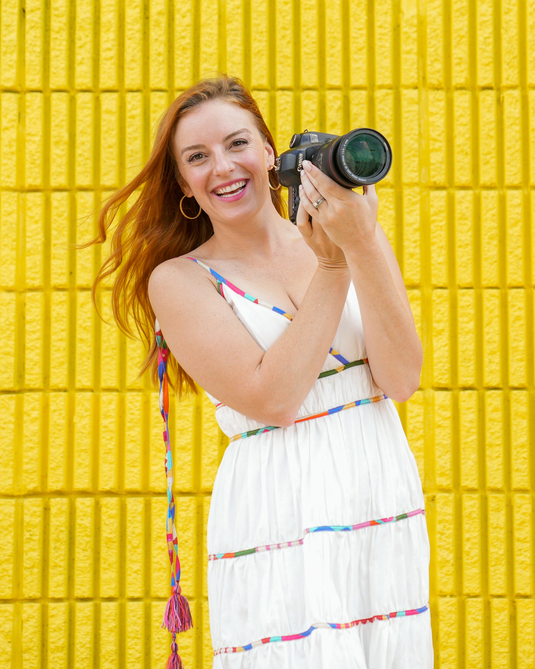 Smiling woman holding a camera against a yellow brick wall.