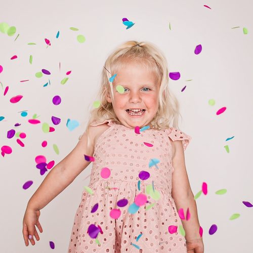 Smiling blonde girl in pink dress with colorful confetti falling around her.