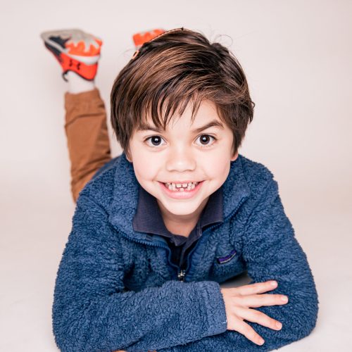 Smiling boy lying down with crossed arms, wearing a blue sweater.